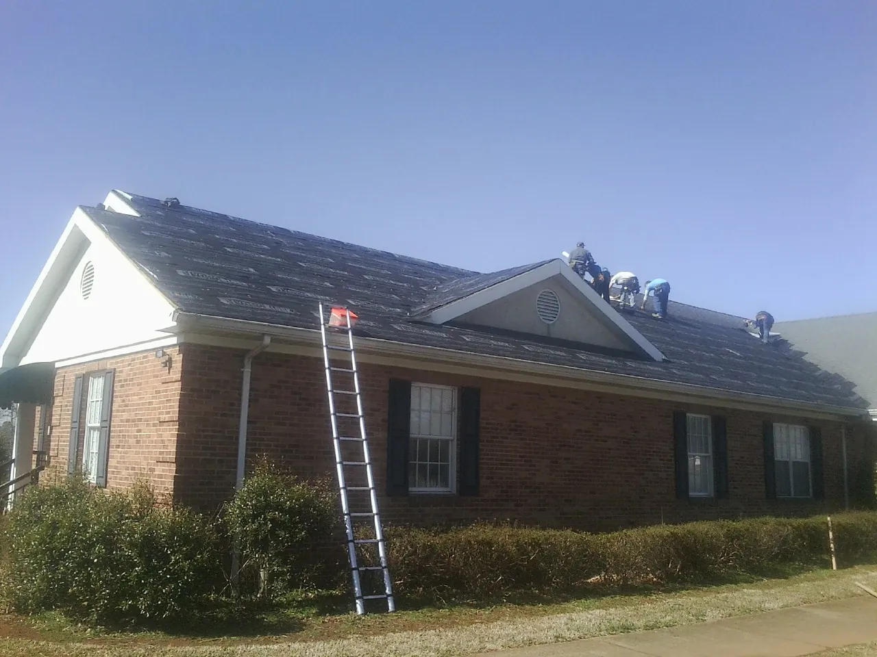 Crew tearing shingles off a brick ranch roof with a ladder set — Emergency Roof Repair work by our crew in College Grove