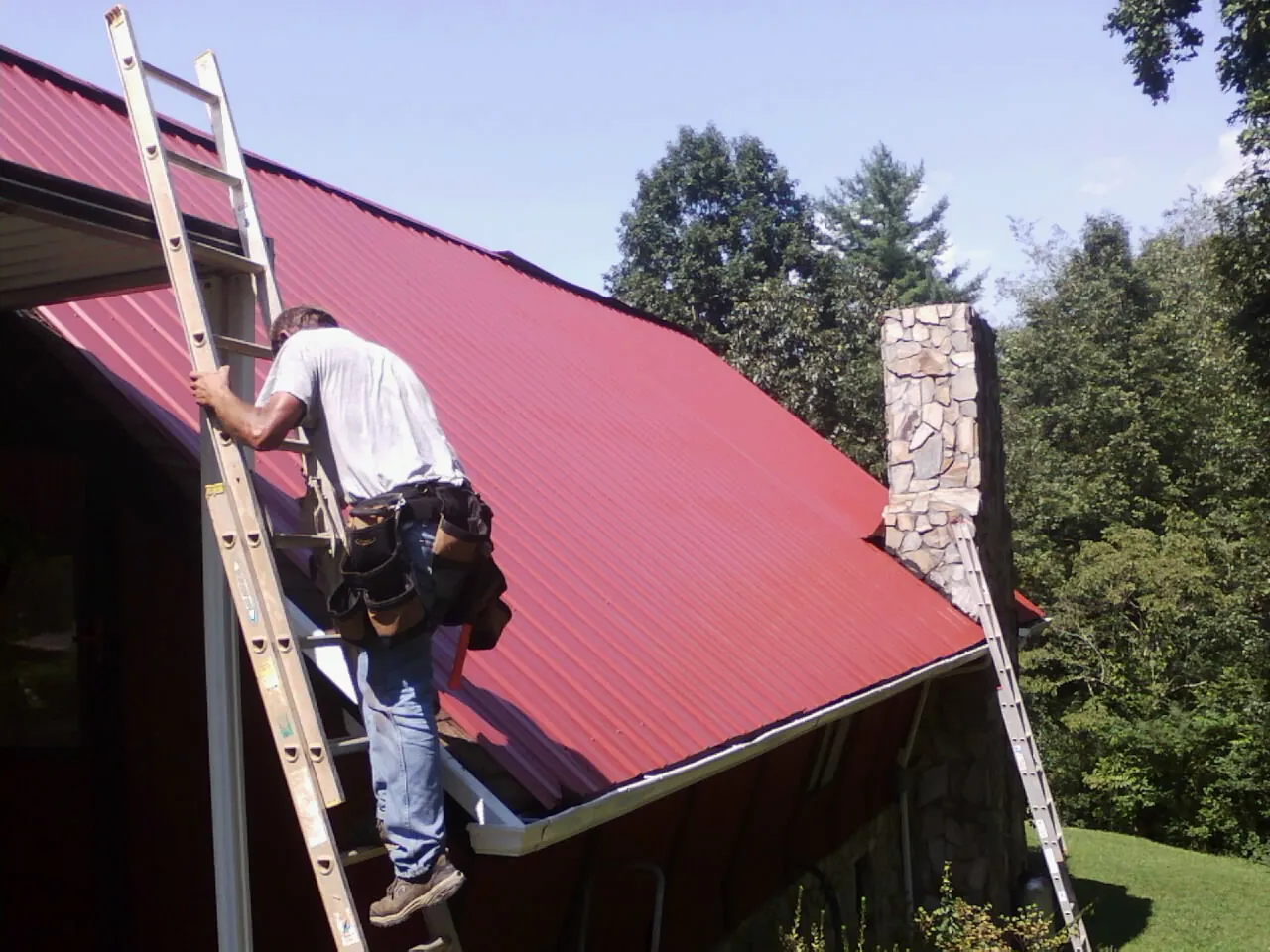 Roofer on a ladder beside a red metal roof and stone chimney — Soffit & Fascia Repair by Roofing Brothers in College Grove